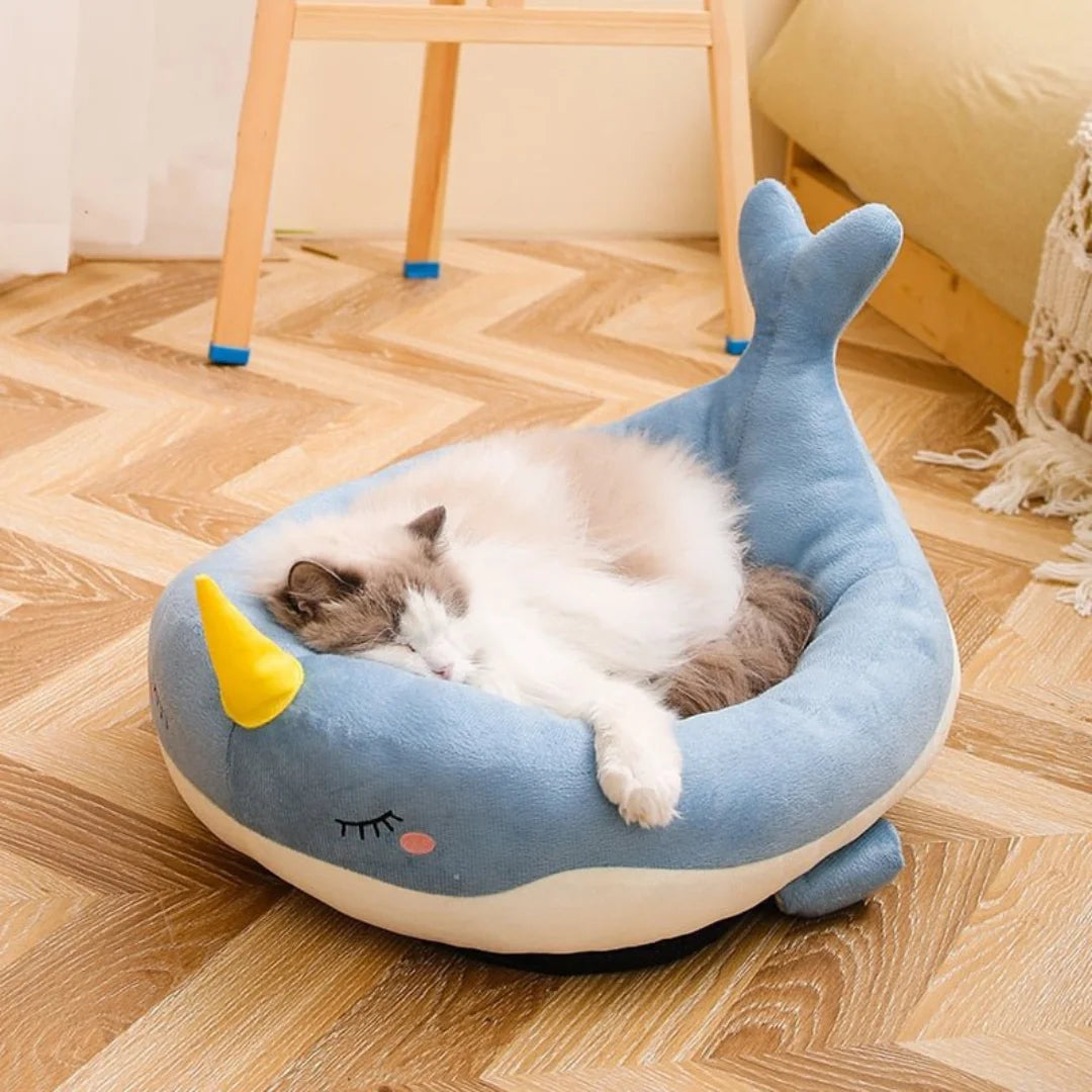 Cat resting comfortably inside the blue narwhal Petifo plush bed on a wooden floor.